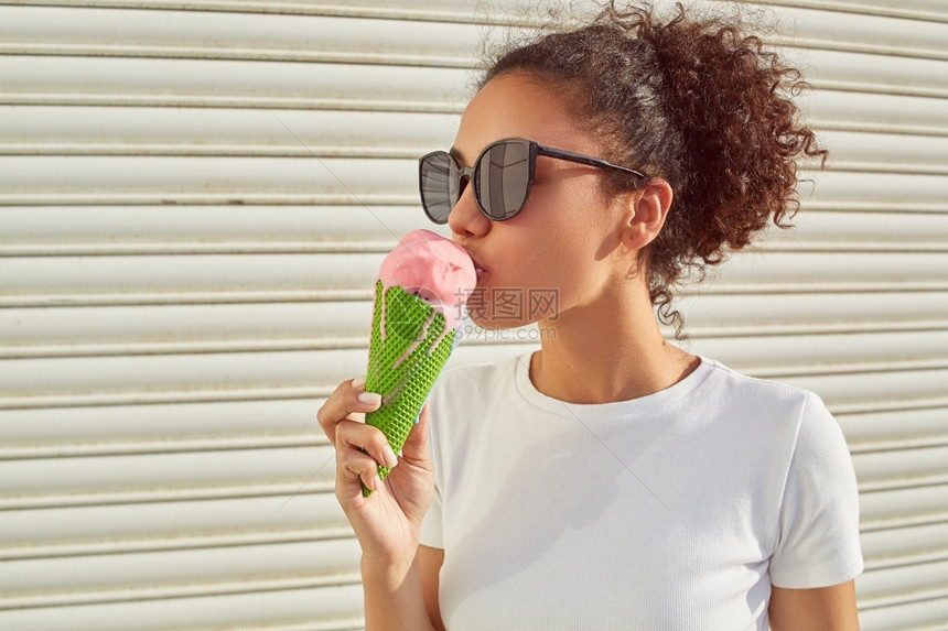 穿着白色T恤和轻牛仔裤的年美籍黑人女孩在日光照耀时用冰淇淋抵挡着一堵轻墙吃冰淇淋白色的模型愉快图片