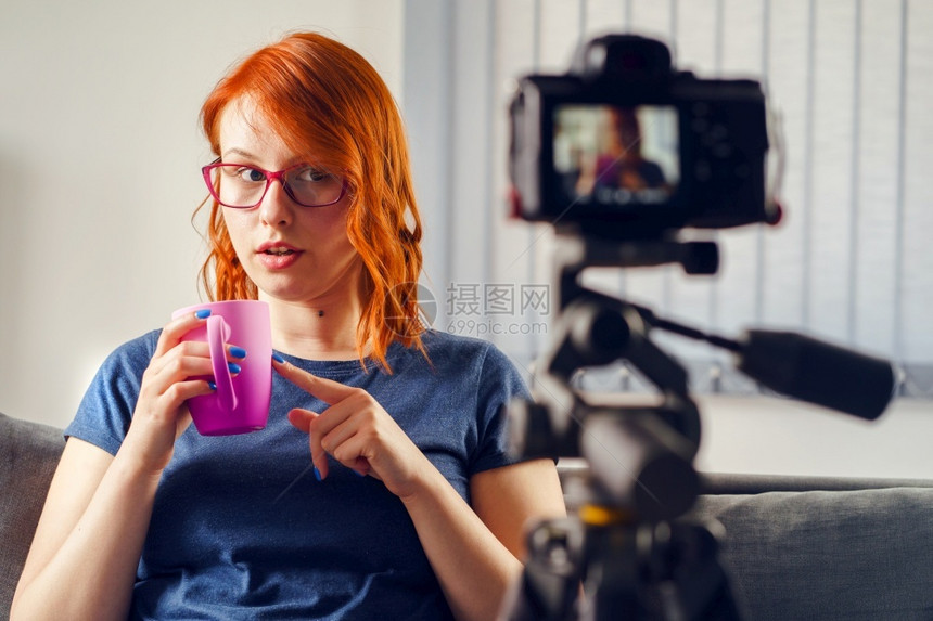 年轻女caucasian女博客在家中或办公室制作视频博客的摄影机前展示茶杯或咖啡穿着T恤衫和眼镜看头前视杯子审查拍摄图片
