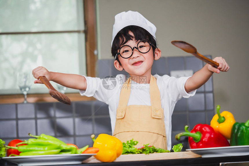 享受亚洲男孩烹饪沙拉食物亚洲男孩烹煮沙拉盛着藏番茄和胡萝卜的蔬菜木勺子铃辣椒为幸福家庭做饭的快乐烤菜亚洲人围裙图片