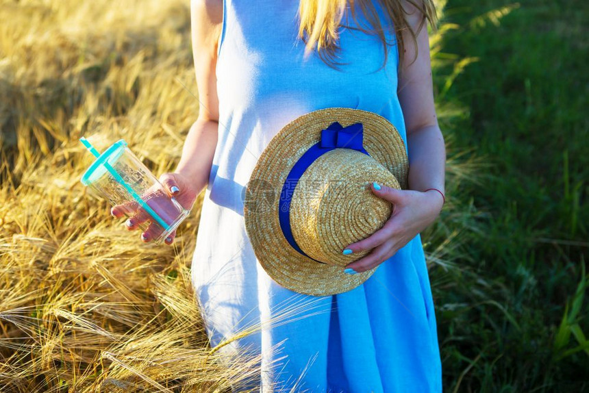 持有穿蓝色裙子的漂亮女孩手里拿着玻璃和帽子穿蓝色裙的漂亮女孩手里拿着玻璃和帽子植物麦田图片