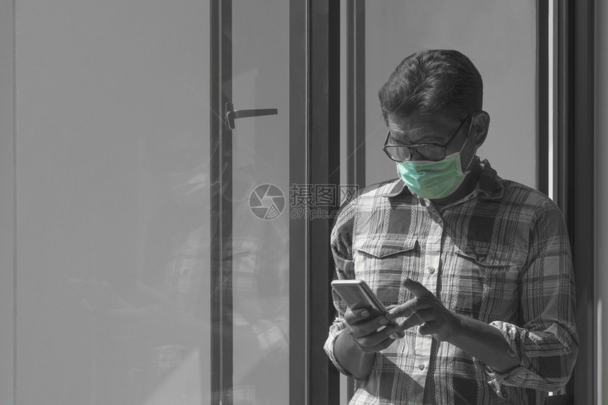 电话格子呢复制身戴保护面罩和使用智能手机的亚裔男子在家客厅玻璃窗附近使用智能手机黑白两色带有绿喷雾技术图片