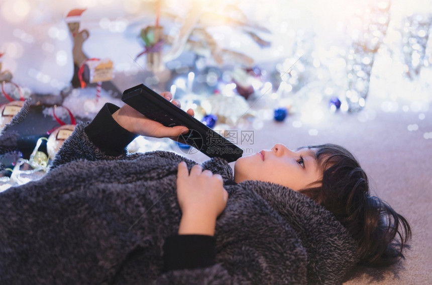 模型内容人们小孩躺在地毯上拿着遥控器坐在地毯上看着电视模糊而明亮的圣诞装饰背景一个快乐的男孩在圣诞节晚上家里放松图片