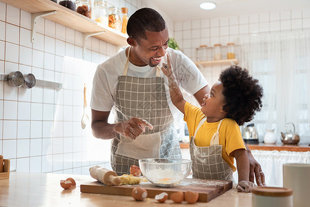 他的面团微笑快乐的黑人儿子在家做面包店时享受玩耍用手指和面粉摸他父亲的鼻子以及指头和面粉在厨房里非洲家庭可以一起在厨房做烘烤蛋糕背景图片