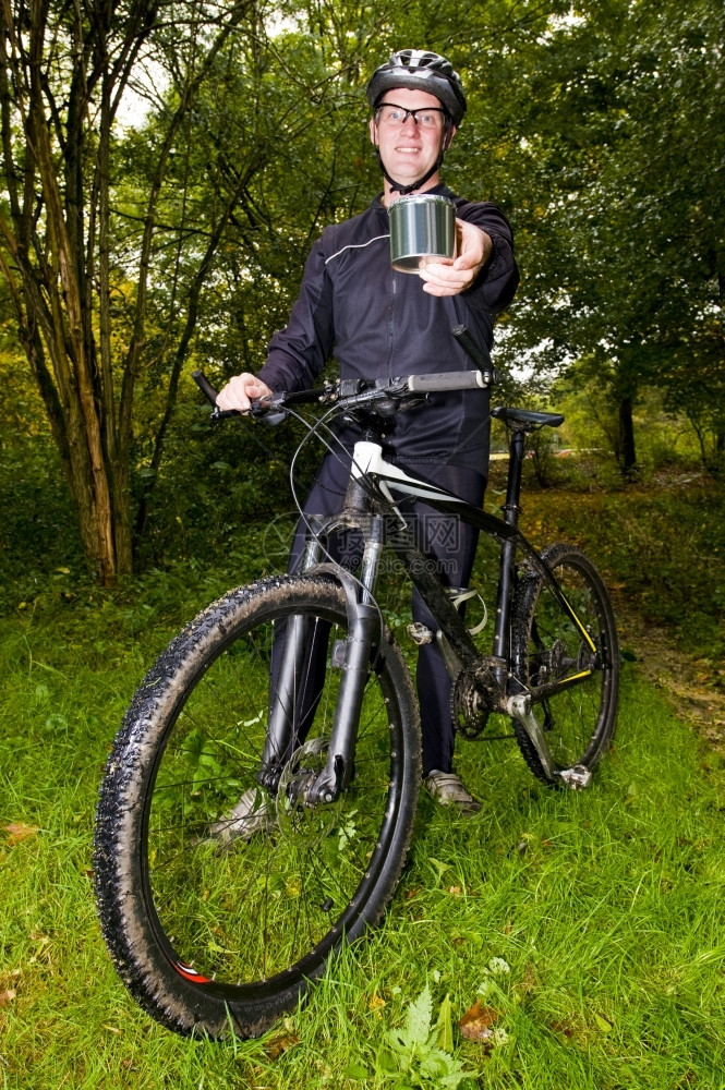 离开链一个笑着的山上骑摩托车者站在他的自行车旁边在他的伸展手举着一个闪亮的钢铁罐路图片