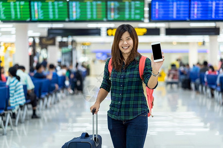 亚洲旅行者携带李在现代机场飞行信息屏幕的带有技术概念的旅行和运输上显示智能移动电话以登入飞行板的亚洲旅者和及运输工具挤移动的亚洲图片