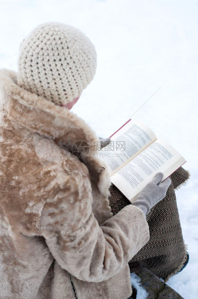 外套户寒冷的一名成年妇女穿着毛皮大衣和羊帽坐在石凳上冬季雪覆盖的公园里阅读一本书图片