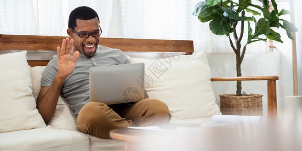 坐在沙发上笔记本与朋友和家人交谈时在笔记本上用打电脑的网上视频话SmingBlack男微笑使用互联网技术享受对话家庭快乐的虚拟的高清图片素材