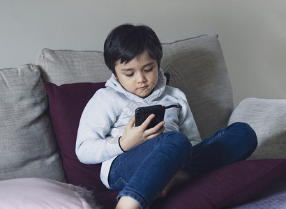 家校互联观看电话坐在沙发上看手机卡通片的男孩在校儿童放学时使用手机在互联网上学习教训的校儿童在家上学的庭就远程习在线教育概念疾病背景