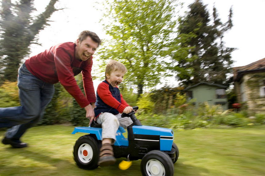 一种乐趣背部父亲把他儿子推在玩具拖拉机上在他们后面的花园主题振奋家庭团结里图片
