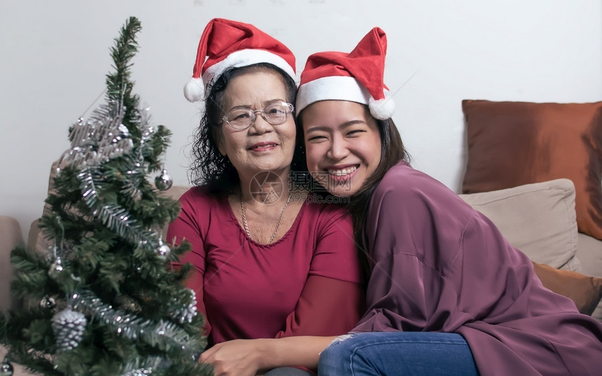 或者高级的两个60岁老母亲和女儿戴着圣塔帽拥抱并坐在家中的客厅里树头模糊圣诞或新年庆典时装瓶子圣诞节或新年庆典概念陈图片