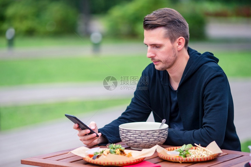 年轻男人在户外咖啡馆吃亚洲街边食物和智能手机工作年轻男人在街上吃东西把面条抢走镇一顿饭旅游图片