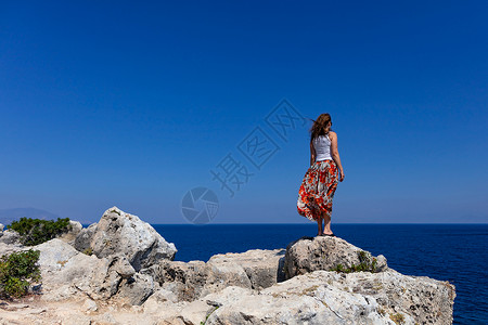 白色上衣搭短裙身穿多彩裙子和白色上衣的年轻女子站在悬崖边缘看着平淡蓝色海面边景岩石缘很多空气有模仿间的图像女士站在石头悬崖的边缘望向海平面的距背景