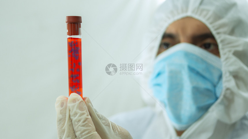 戴着面具和白色生物防护服的医拿着一个带有Covid19标记的试管冠状血液样本用手漂浮在试管内并白色背景上专心地盯着它血样用手漂浮图片