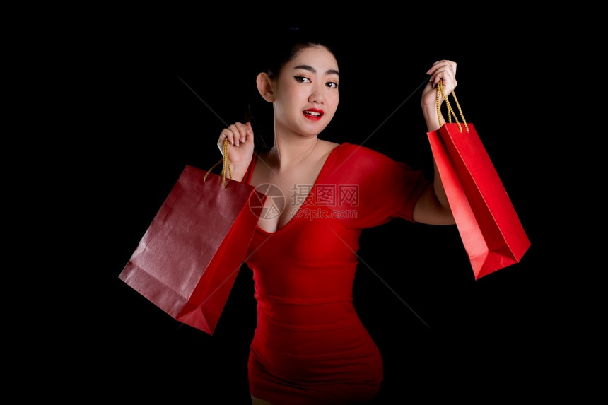 一位美丽的亚洲年轻女身着红色礼服持有信用卡和购物袋的黑背景销售概念照相机等被描绘成肖像模型兴奋的时尚图片
