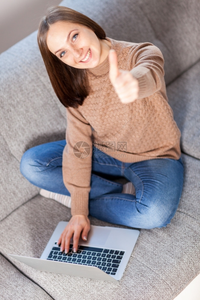 照片中笑着的caucasian女人在用电脑坐沙发上时做积极的拇指手势学生年轻的尽管图片