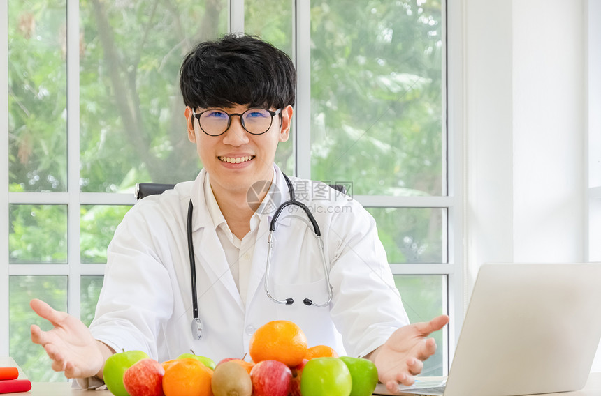 保持关心吃微笑的年男营养学家戴着身穿实验室大衣制服的眼镜在办公室桌子上看着照相机将健康的新鲜苹果橙子和kiwifruit鲜果子放图片