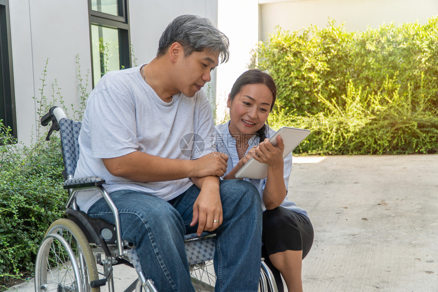 医院爱坐在轮椅上妻子坐在旁边并拿着平板电脑病人因车祸而坐轮椅妻子在旁边让他花园漫步时放松一下不要让他看到与肩并和鼓励的观念药片图片