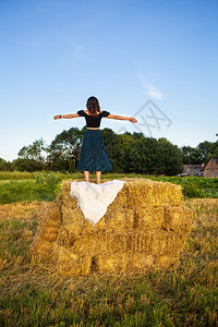 大包干草年轻美女站在一大堆稻草捆上自由风落日年轻美女站在一大堆稻草捆上自由落日夏天肩膀背景