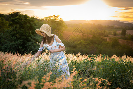 太阳裙假期女孩戴帽子穿白色衣服站在草地中间穿着美丽的白花在太阳下定时间在山上心情放松和快乐的山上她戴着帽子穿白色裙站在草丛中温暖的花朵背景
