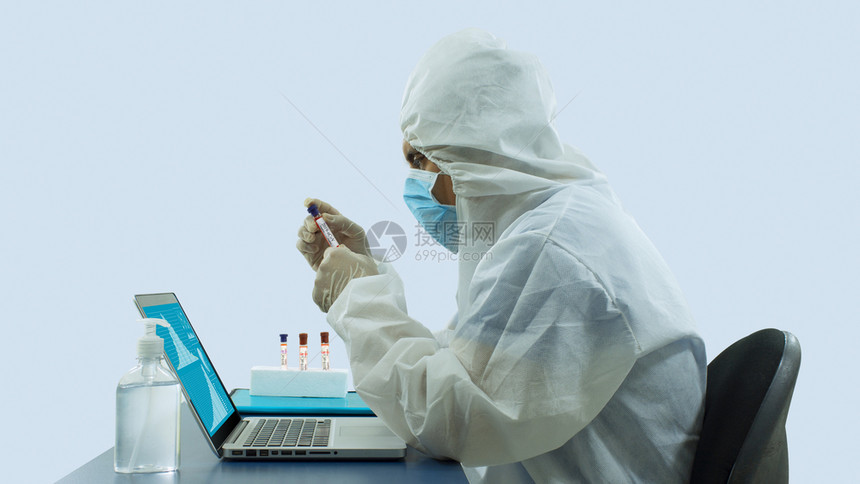 保持服装戴着蓝色面罩和生物防护服的医在办公室里用笔记本电脑工作的侧视图手里拿着一个试管注意力集中白色背景的桌子上放着一瓶酒精凝胶图片