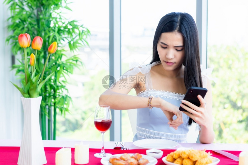 年轻的可爱情人节概念亚洲年轻女孩坐在餐桌食物旁用酒杯和红玫瑰粉的花束看手表在餐馆等她的男人在餐厅里等待她的男人晚餐图片