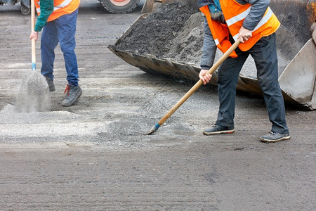 巷道工作灰色的身穿明橙反射背心的运输工人在清理公路上的旧沥青之后为修车准备旧路面并将道残骸清除成一个平板桶然后从公路上清除旧沥青背景