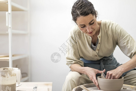 慢的陶瓷制品工作室年轻女子手持陶器刮磨塑女士坐在长凳上用陶轮做罐图片