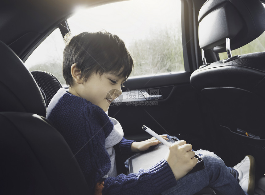 快乐的年轻男孩坐在带有安全的汽车后座乘客位上时使用平板电脑男孩子在智能垫上画窗户里面活动图片
