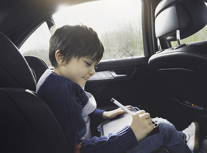 车后座位快乐的年轻男孩坐在带有安全的汽车后座乘客位上时使用平板电脑男孩子在智能垫上画窗户里面活动背景