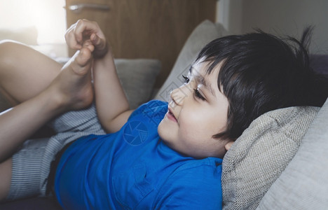 快乐的健康杰米坐在沙发上躺脚指钉着甲看电视在家放松快乐儿童室内肖像男孩在家中画像男孩新式正常生活方图片
