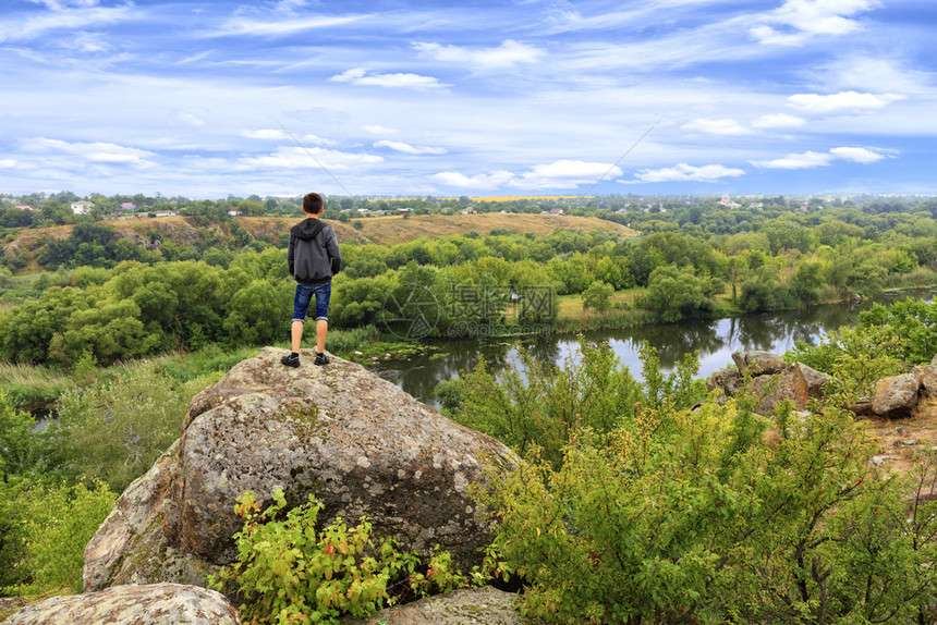 河床一个十几岁的少年站在南虫河岸的巨石堆上看着地平线夏天的南虫河是岩石般的海岸明亮绿植被和一片阴暗的蓝天A青少年站在南虫河岸的巨图片