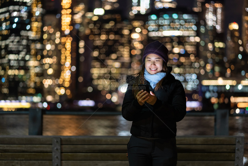 天际线女商人穿着冬季西装的亚洲女使用智能手机与微笑动作在东河背景美国市中心的纽约城风景照片上模糊的相布基赫Bokoh旁边人们图片