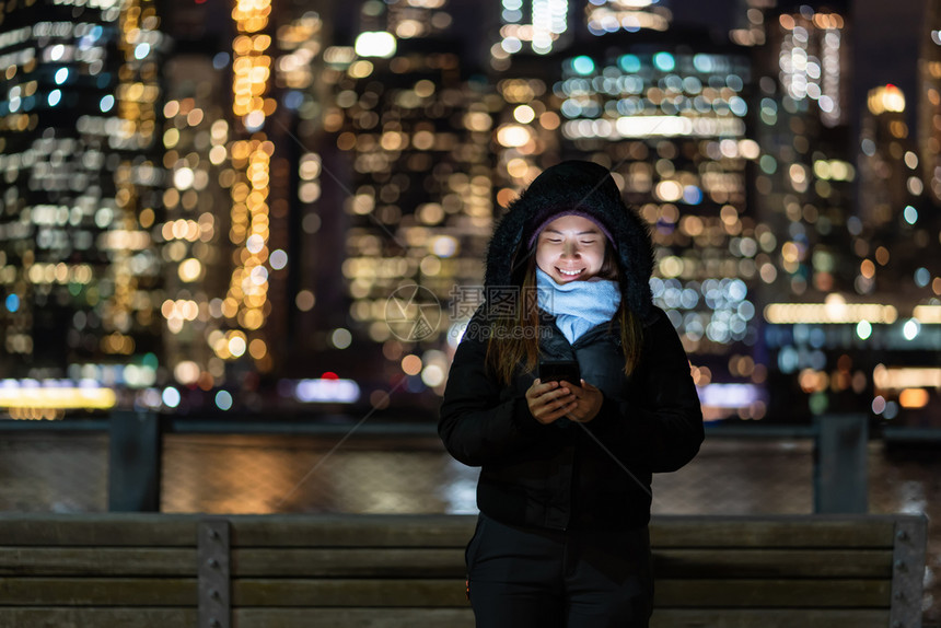 夜晚专业的穿着冬季西装亚洲女使用智能手机与微笑动作在东河背景美国市中心的纽约城风景照片上模糊的相布基赫Bokoh旁边自由职业者图片