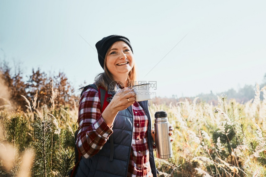 微笑的女人在假期旅行中放松和享受咖啡女人站在小路上看着远方手里拿一杯咖啡和保温瓶旅行女人站在小路上望着远方手里拿一杯咖啡和保温瓶图片