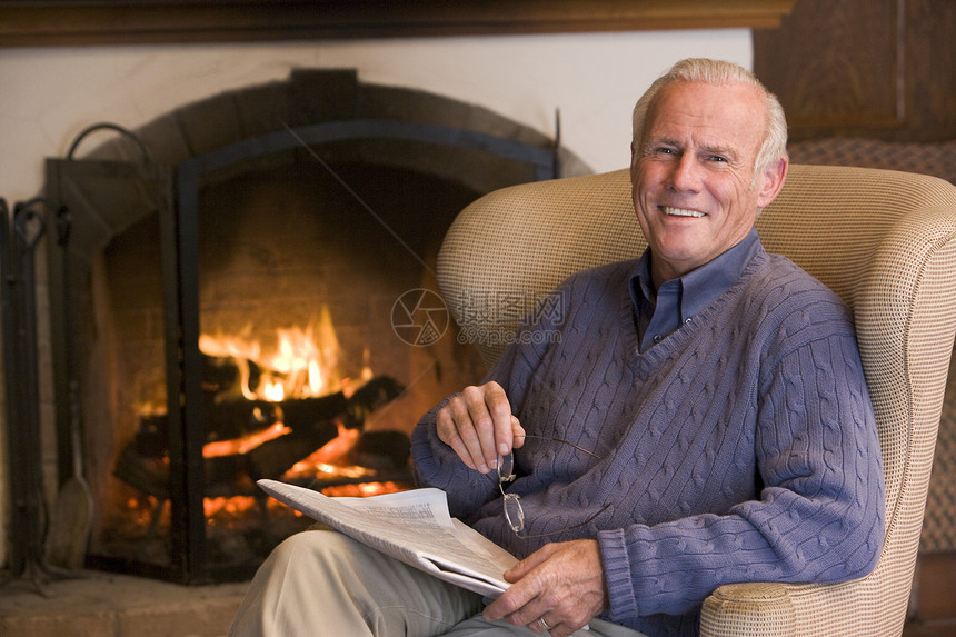 坐在壁炉边起居室的男子 报纸微笑着男人水平放松相机长度退休木火时事男性椅子图片