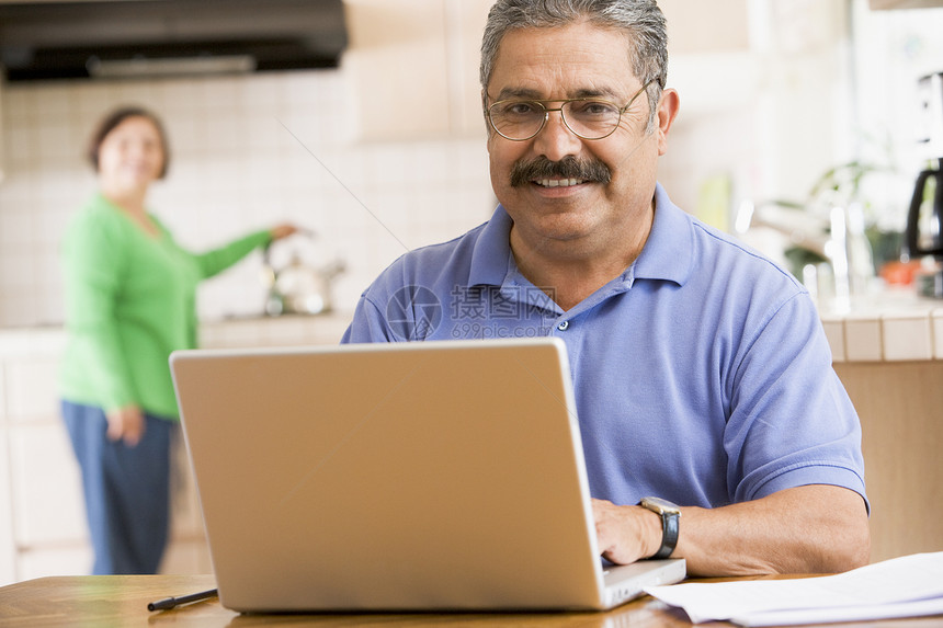 在厨房里的男人 手持笔记本电脑的人 与背景中的女人微笑成人夫妻互联网冲浪婴儿中年技术拉丁银色女士图片