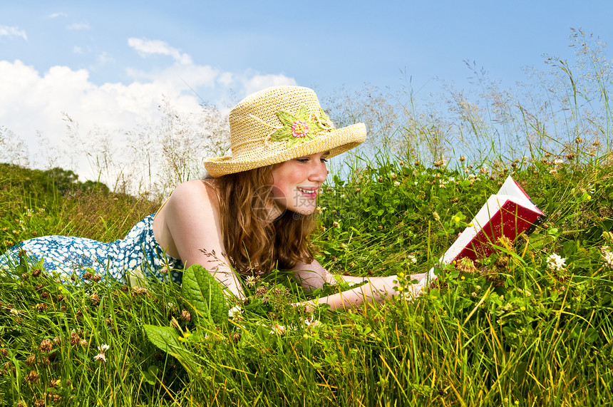 年轻女孩在草原读书青少年微笑蓝色女孩们草地享受精装草帽稻草晴天图片