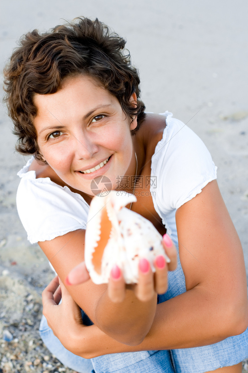女人在海边海岸 用一只壳手握着享受旅行头发假期黑发牛仔裤成人深色天空蓝色图片
