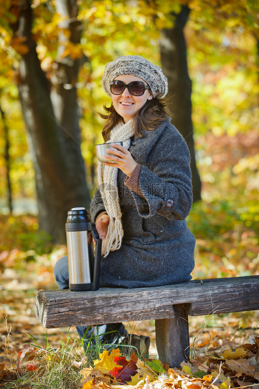 女人在秋天公园喝茶热水瓶乐趣眼镜杯子女孩太阳微笑季节女性青年图片