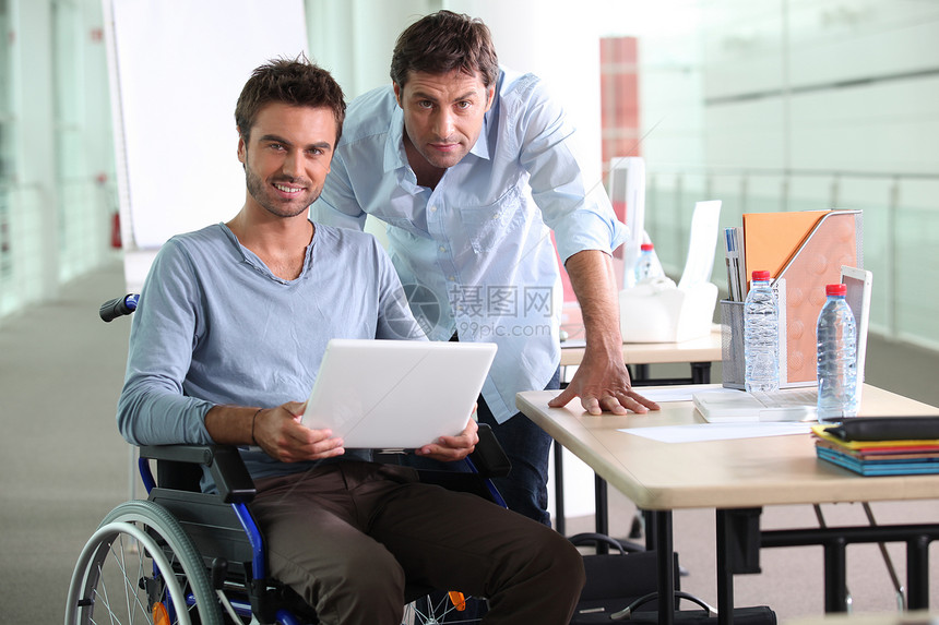 男人坐在轮椅上 手持笔记本电脑与同事并肩工作图片