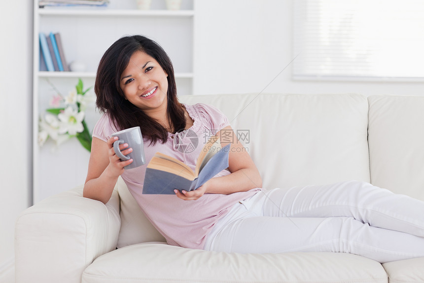 躺在沙发上时 微笑的女人拿着一本书和一张杯子图片
