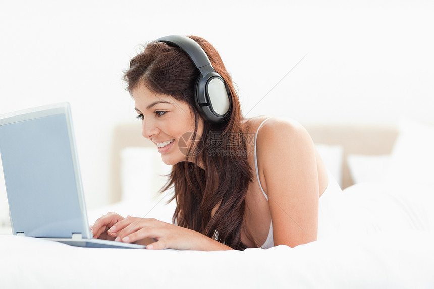 妇女躺在床上时用耳机浏览笔记本电脑图片