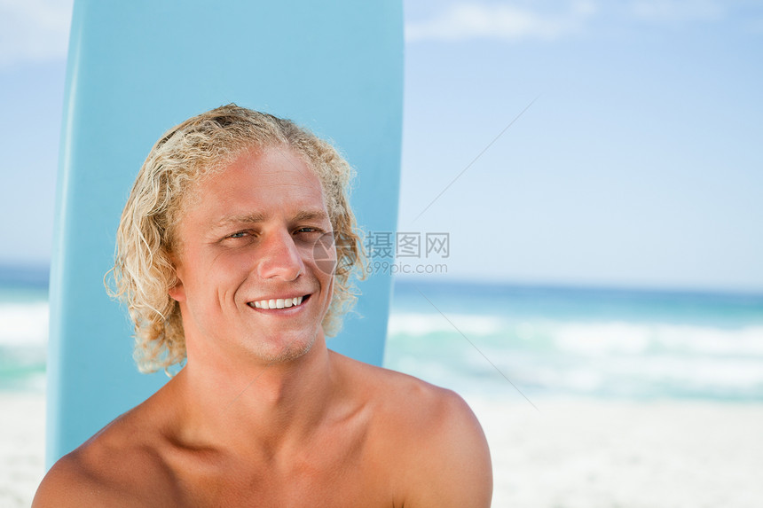 坐在海洋前面的微笑的年轻人 用他的冲浪板笑图片
