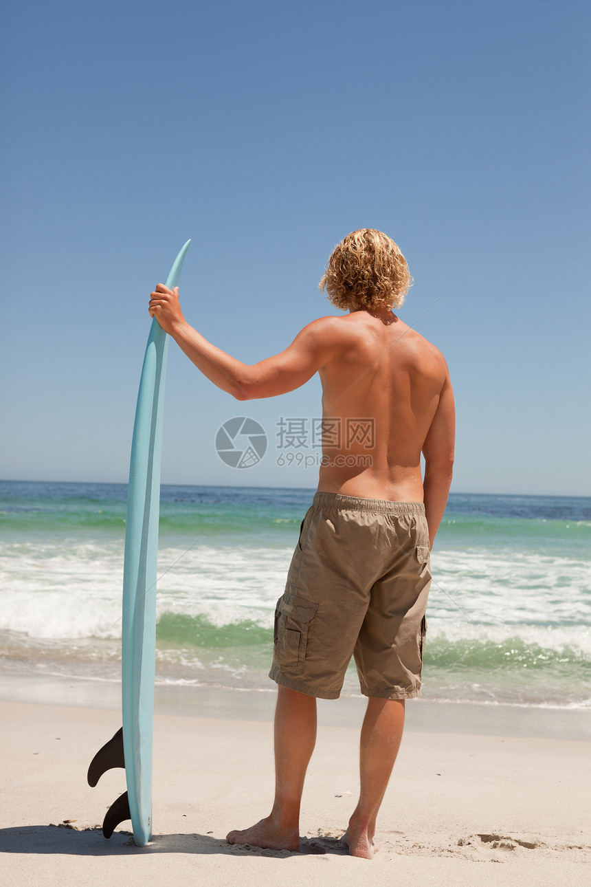 年轻的金发金发男 在海洋前拿着他的冲浪板图片