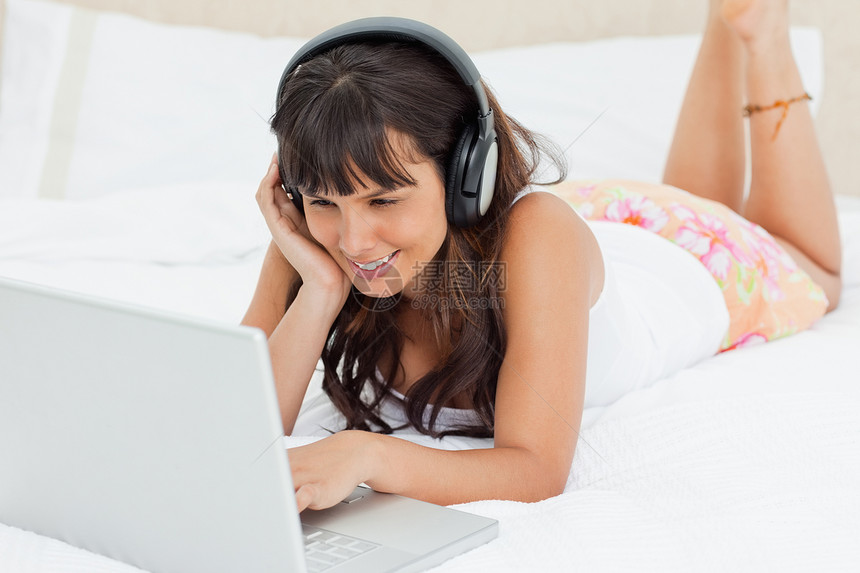 微笑的年轻女子观看录像学生卧室笔记本睡衣腹部电脑外表耳机白色女性图片