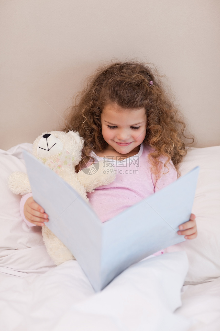微笑的女孩在读一本书拥抱外表阅读玩具熊孩子枕头女孩们毯子就寝寝具图片