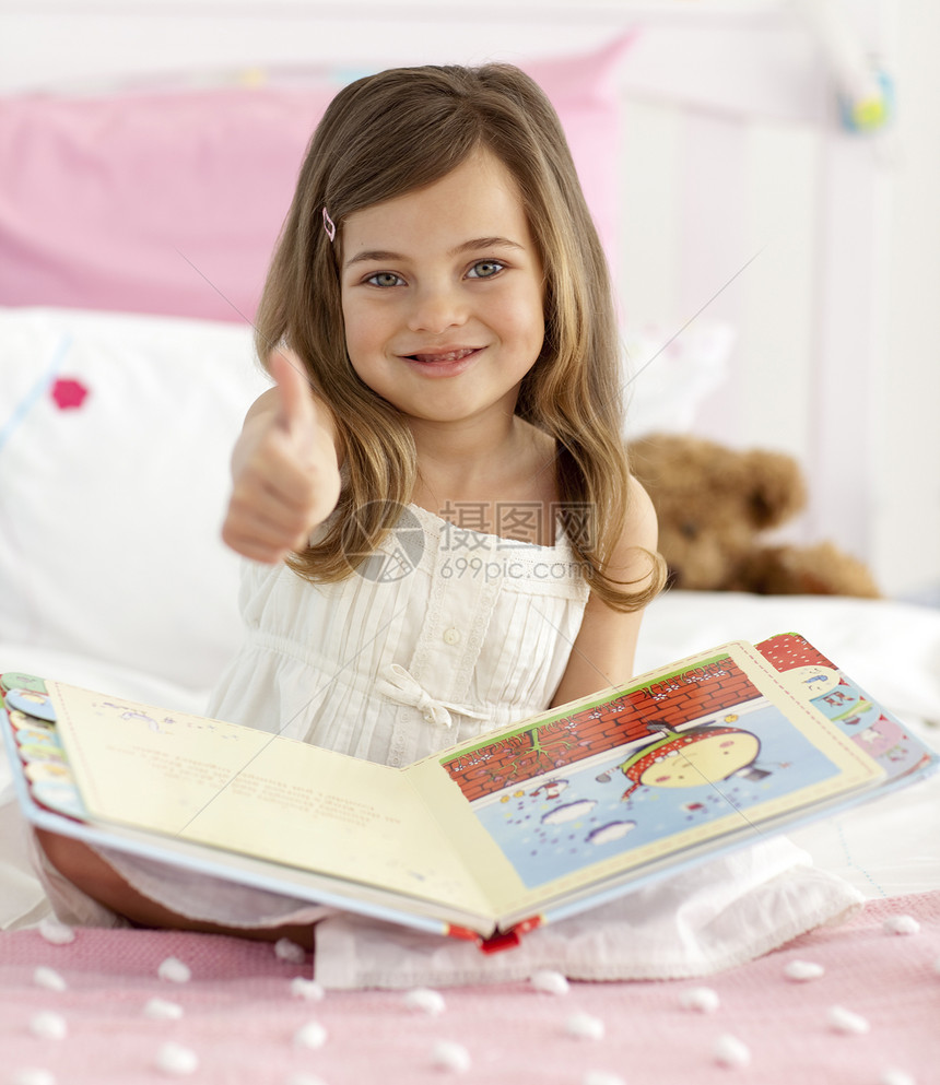 快乐的小女孩在床上看书时举起拇指图片