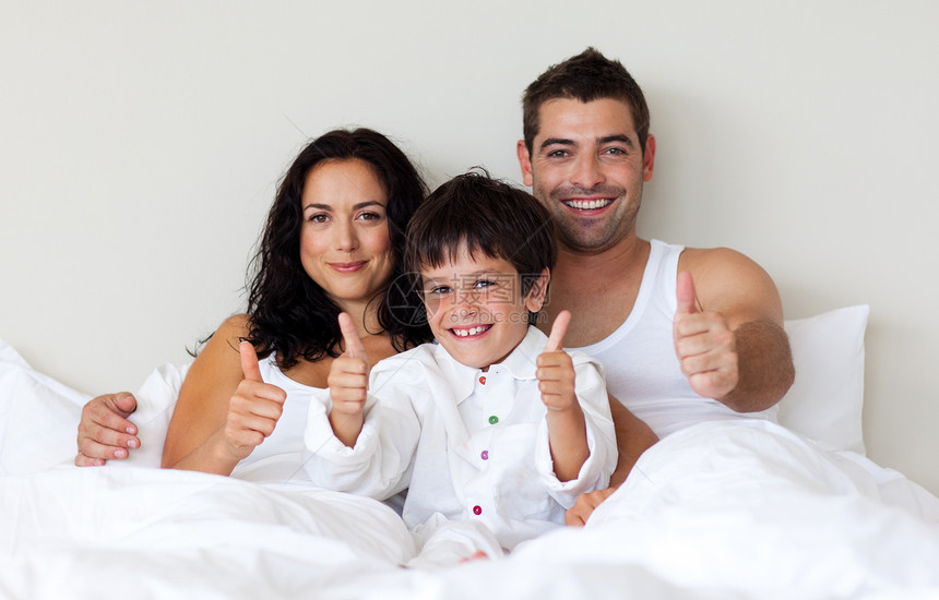 快乐的儿子 用拇指抬起他的大拇指 和他的父母在床上图片