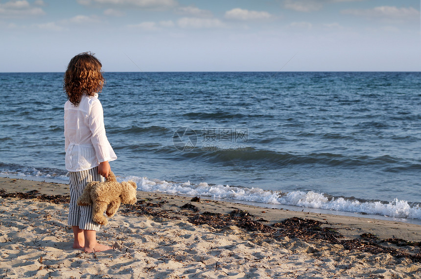 带着泰迪熊的小女孩 站在海滩上 看着图片