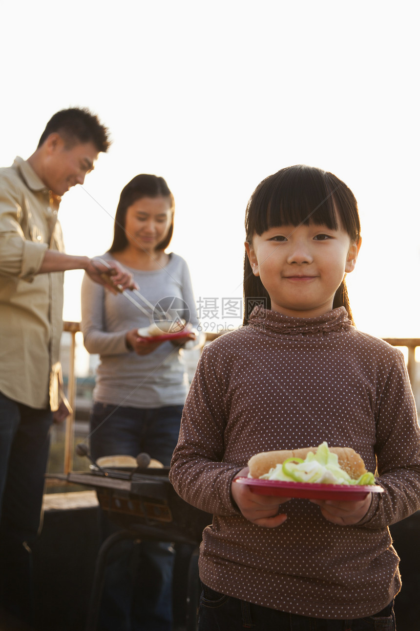 女儿拿着热狗和沙拉的盘子 父母站在烤肉旁边图片